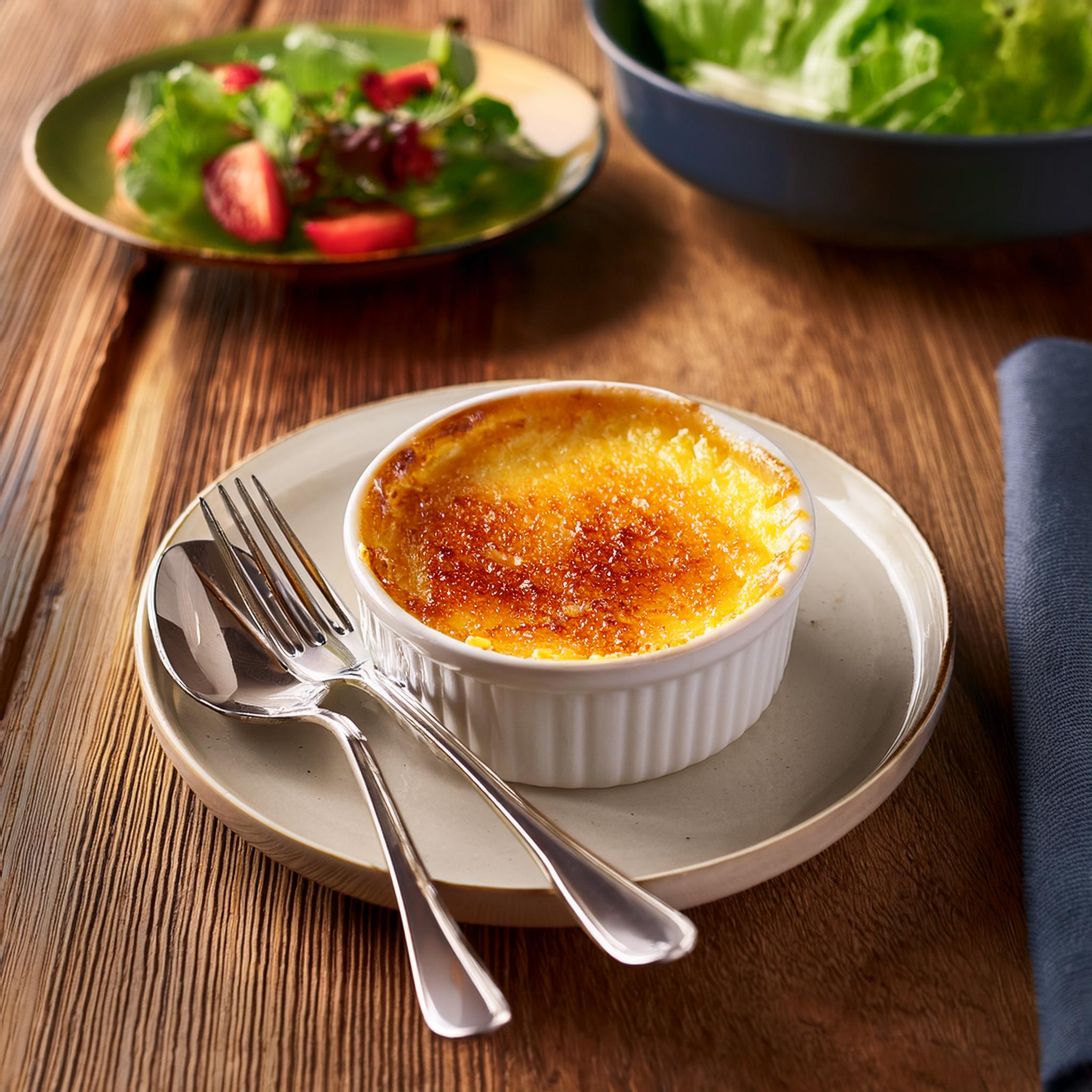 A gourmet Crab Brulee dish, beautifully plated in a small ramekin with a golden, caramelized sugar crust on top. The dish is presented on a rustic wooden table with elegant cutlery and a light salad on the side. Soft lighting highlights the glossy, crispy top and the creamy custard underneath.